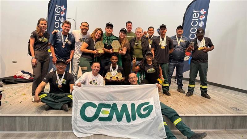 A imagem mostra um grupo de pessoas reunidas em um palco, todas vestindo uniformes e exibindo medalhas. Alguns seguram troféus, o que indica que participaram de uma competição ou cerimônia de premiação. Na frente, duas pessoas seguram uma faixa branca com o logotipo verde e amarelo da CEMIG. Ao fundo, há dois banners verticais com a palavra Cacier e outros logotipos. O ambiente é claramente de um evento formal, voltado para reconhecimento e celebração de conquistas coletivas.