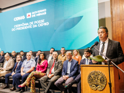 A imagem mostra uma cerimônia oficial em Minas Gerais, Brasil. No palco, há várias pessoas sentadas em cadeiras, enquanto uma pessoa está em pé atrás de um púlpito, fazendo um discurso. O púlpito traz o brasão do estado de Minas Gerais. Ao fundo, um grande painel azul-esverdeado exibe os dizeres: “CEMIG – Governo de Minas – Cerimônia de Posse do Presidente da CEMIG”, indicando que se trata da solenidade de posse do novo presidente da companhia energética CEMIG. O ambiente é formal, com autoridades e convidados presentes, reforçando o caráter institucional e governamental do evento.