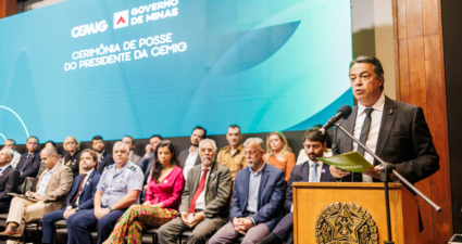 A imagem mostra uma cerimônia oficial em Minas Gerais, Brasil. No palco, há várias pessoas sentadas em cadeiras, enquanto uma pessoa está em pé atrás de um púlpito, fazendo um discurso. O púlpito traz o brasão do estado de Minas Gerais. Ao fundo, um grande painel azul-esverdeado exibe os dizeres: “CEMIG – Governo de Minas – Cerimônia de Posse do Presidente da CEMIG”, indicando que se trata da solenidade de posse do novo presidente da companhia energética CEMIG. O ambiente é formal, com autoridades e convidados presentes, reforçando o caráter institucional e governamental do evento.