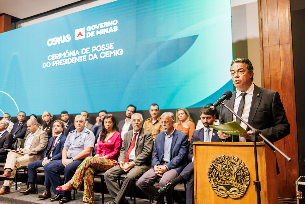 A imagem mostra uma cerimônia oficial em Minas Gerais, Brasil. No palco, há várias pessoas sentadas em cadeiras, enquanto uma pessoa está em pé atrás de um púlpito, fazendo um discurso. O púlpito traz o brasão do estado de Minas Gerais.

Ao fundo, um grande painel azul-esverdeado exibe os dizeres: “CEMIG – Governo de Minas – Cerimônia de Posse do Presidente da CEMIG”, indicando que se trata da solenidade de posse do novo presidente da companhia energética CEMIG. O ambiente é formal, com autoridades e convidados presentes, reforçando o caráter institucional e governamental do evento.