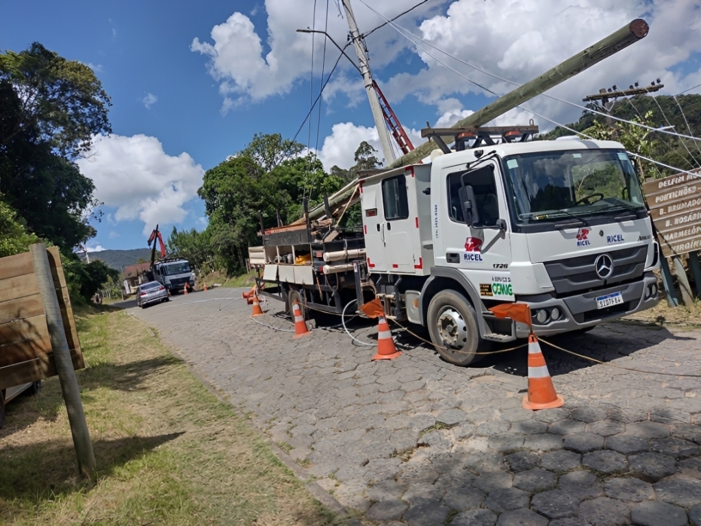 A imagem mostra uma operação de manutenção de infraestrutura elétrica em uma estrada de paralelepípedos. Um caminhão utilitário identificado com as marcas “RICEL” e “CEMIG” está estacionado, cercado por cones laranja e cabos, e equipado com um braço mecânico que ergue ou posiciona um poste de madeira — provavelmente para instalação ou reparo de linhas de energia ou telefonia.

Ao fundo, há outro caminhão e um carro, enquanto a estrada é ladeada por árvores e vegetação. À direita, um poste de madeira traz placas indicando direções para localidades próximas, como “DELFIN MOREIRA”, “POTENTINHOS”, “B. BRUMADÃO”, “B. ROSÁRIO”, “B. NAZAREMÓ” e “VIRGOLÂNDIA”.

A cena transmite claramente o trabalho de manutenção elétrica, envolvendo infraestrutura pública e segurança viária, mostrando como equipes técnicas atuam em campo para garantir serviços essenciais às comunidades.