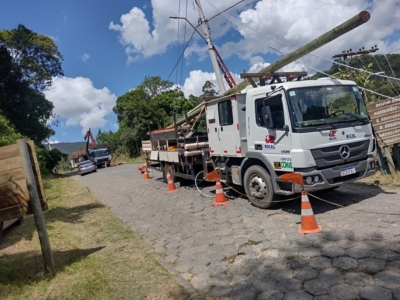A imagem mostra uma operação de manutenção de infraestrutura elétrica em uma estrada de paralelepípedos. Um caminhão utilitário identificado com as marcas “RICEL” e “CEMIG” está estacionado, cercado por cones laranja e cabos, e equipado com um braço mecânico que ergue ou posiciona um poste de madeira — provavelmente para instalação ou reparo de linhas de energia ou telefonia. Ao fundo, há outro caminhão e um carro, enquanto a estrada é ladeada por árvores e vegetação. À direita, um poste de madeira traz placas indicando direções para localidades próximas, como “DELFIN MOREIRA”, “POTENTINHOS”, “B. BRUMADÃO”, “B. ROSÁRIO”, “B. NAZAREMÓ” e “VIRGOLÂNDIA”. A cena transmite claramente o trabalho de manutenção elétrica, envolvendo infraestrutura pública e segurança viária, mostrando como equipes técnicas atuam em campo para garantir serviços essenciais às comunidades. Legenda: A dupla alimentação interliga as redes de distribuição a dois circuitos independentes, possibilitando o redirecionamento do fornecimento de energia em casos de falha ou necessidade de manutenção em um dos alimentadores