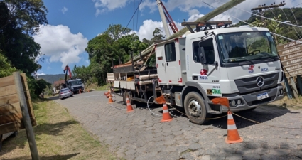 A imagem mostra uma operação de manutenção de infraestrutura elétrica em uma estrada de paralelepípedos. Um caminhão utilitário identificado com as marcas “RICEL” e “CEMIG” está estacionado, cercado por cones laranja e cabos, e equipado com um braço mecânico que ergue ou posiciona um poste de madeira — provavelmente para instalação ou reparo de linhas de energia ou telefonia. Ao fundo, há outro caminhão e um carro, enquanto a estrada é ladeada por árvores e vegetação. À direita, um poste de madeira traz placas indicando direções para localidades próximas, como “DELFIN MOREIRA”, “POTENTINHOS”, “B. BRUMADÃO”, “B. ROSÁRIO”, “B. NAZAREMÓ” e “VIRGOLÂNDIA”. A cena transmite claramente o trabalho de manutenção elétrica, envolvendo infraestrutura pública e segurança viária, mostrando como equipes técnicas atuam em campo para garantir serviços essenciais às comunidades. Legenda: A dupla alimentação interliga as redes de distribuição a dois circuitos independentes, possibilitando o redirecionamento do fornecimento de energia em casos de falha ou necessidade de manutenção em um dos alimentadores
