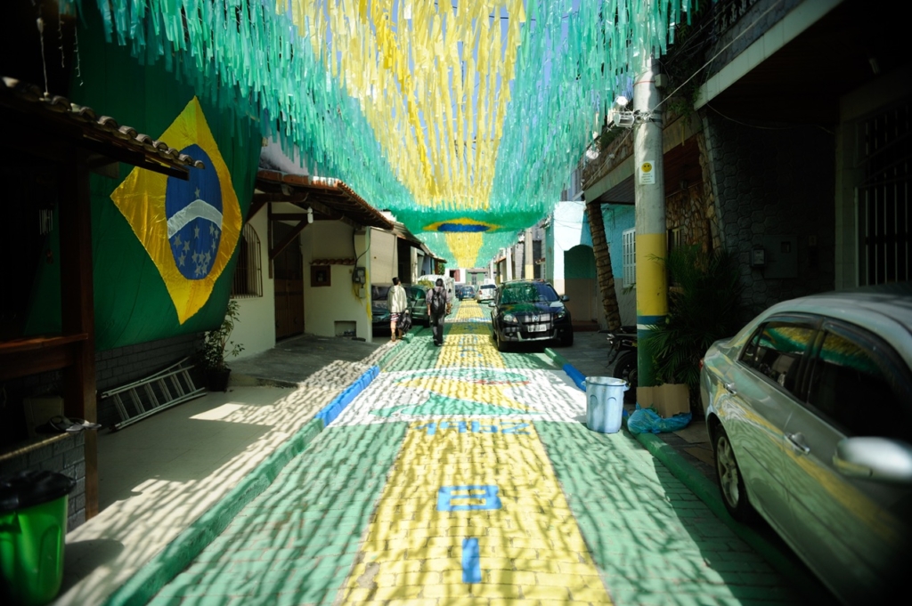 A imagem mostra uma rua residencial estreita, toda decorada com as cores da bandeira do Brasil — verde, amarelo e azul. O chão foi pintado com um grande desenho da bandeira brasileira, criando um efeito vibrante. No alto, faixas longas de tecido verde e amarelo atravessam a rua, formando uma espécie de cobertura festiva. À esquerda, na parede de uma casa, há uma bandeira do Brasil pendurada em destaque. Alguns carros estão estacionados ao longo da rua e duas pessoas caminham tranquilamente pelo cenário. O conjunto transmite um forte espírito comunitário e nacional, provavelmente ligado a uma celebração como um evento esportivo ou uma data cívica.

Esse tipo de decoração é bastante comum em épocas de Copa do Mundo ou festas juninas, quando os moradores se unem para transformar o bairro em um espaço de celebração. 