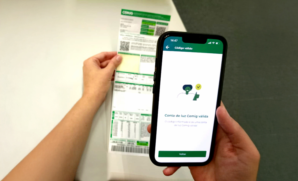 A imagem mostra uma cena cotidiana de verificação de contas de energia elétrica. Uma pessoa segura um smartphone em uma mão e uma conta de luz da Cemig (Companhia Energética de Minas Gerais) na outra.
Na tela do celular aparece uma mensagem em português confirmando que o código informado é válido:
“Código válido
Conta de luz Cemig válida
O código informado é de uma conta de luz Cemig válida!”
Logo abaixo, há um botão verde escrito “Voltar”.
A conta impressa traz o logotipo da Cemig no topo, além de tabelas, códigos de barras e números relacionados ao consumo e ao valor da fatura. A cena transmite a ideia de integração digital, mostrando como é possível validar informações da conta de energia diretamente pelo aplicativo no celular, sem depender apenas do papel.
Esse tipo de prática reflete o avanço da gestão digital de contas e a conveniência oferecida pelas empresas de serviços públicos.