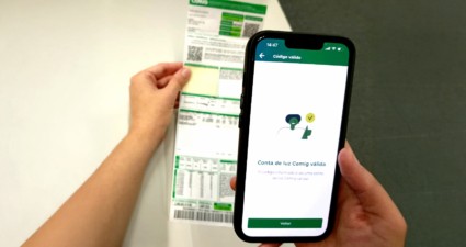 A imagem mostra uma cena cotidiana de verificação de contas de energia elétrica. Uma pessoa segura um smartphone em uma mão e uma conta de luz da Cemig (Companhia Energética de Minas Gerais) na outra. Na tela do celular aparece uma mensagem em português confirmando que o código informado é válido: “Código válido Conta de luz Cemig válida O código informado é de uma conta de luz Cemig válida!” Logo abaixo, há um botão verde escrito “Voltar”. A conta impressa traz o logotipo da Cemig no topo, além de tabelas, códigos de barras e números relacionados ao consumo e ao valor da fatura. A cena transmite a ideia de integração digital, mostrando como é possível validar informações da conta de energia diretamente pelo aplicativo no celular, sem depender apenas do papel. Esse tipo de prática reflete o avanço da gestão digital de contas e a conveniência oferecida pelas empresas de serviços públicos.