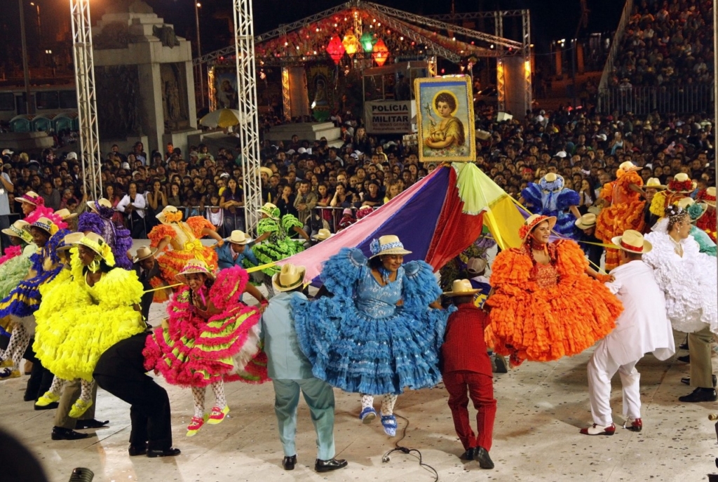 A imagem mostra um festival cultural noturno cheio de energia e tradição. No centro, um grande grupo de dançarinos se apresenta em formação circular, vestidos com trajes folclóricos vibrantes — vestidos rodados com babados em cores vivas como amarelo, rosa, azul, verde e laranja, além de chapéus de aba larga. O cenário transmite alegria e movimento, reforçado pelas lanternas iluminadas e bandeiras festivas que decoram o espaço.
Ao fundo, uma multidão acompanha atentamente a apresentação, enquanto um quadro religioso é erguido acima dos dançarinos, sugerindo que o evento tem também caráter espiritual. A presença de uma faixa com “POLICIA MILITAR” indica que há segurança organizada para garantir a tranquilidade da celebração.
É uma cena que combina tradição popular, dança folclórica e espírito comunitário, transmitindo a riqueza cultural e a força coletiva de uma festa típica brasileira.
