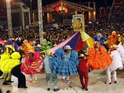 Crédito das fotos: Agência Minas/Robson Vasconcellos A imagem mostra um festival cultural noturno cheio de energia e tradição. No centro, um grande grupo de dançarinos se apresenta em formação circular, vestidos com trajes folclóricos vibrantes — vestidos rodados com babados em cores vivas como amarelo, rosa, azul, verde e laranja, além de chapéus de aba larga. O cenário transmite alegria e movimento, reforçado pelas lanternas iluminadas e bandeiras festivas que decoram o espaço. Ao fundo, uma multidão acompanha atentamente a apresentação, enquanto um quadro religioso é erguido acima dos dançarinos, sugerindo que o evento tem também caráter espiritual. A presença de uma faixa com “POLICIA MILITAR” indica que há segurança organizada para garantir a tranquilidade da celebração. É uma cena que combina tradição popular, dança folclórica e espírito comunitário, transmitindo a riqueza cultural e a força coletiva de uma festa típica brasileira.