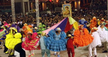 Crédito das fotos: Agência Minas/Robson Vasconcellos A imagem mostra um festival cultural noturno cheio de energia e tradição. No centro, um grande grupo de dançarinos se apresenta em formação circular, vestidos com trajes folclóricos vibrantes — vestidos rodados com babados em cores vivas como amarelo, rosa, azul, verde e laranja, além de chapéus de aba larga. O cenário transmite alegria e movimento, reforçado pelas lanternas iluminadas e bandeiras festivas que decoram o espaço. Ao fundo, uma multidão acompanha atentamente a apresentação, enquanto um quadro religioso é erguido acima dos dançarinos, sugerindo que o evento tem também caráter espiritual. A presença de uma faixa com “POLICIA MILITAR” indica que há segurança organizada para garantir a tranquilidade da celebração. É uma cena que combina tradição popular, dança folclórica e espírito comunitário, transmitindo a riqueza cultural e a força coletiva de uma festa típica brasileira.
