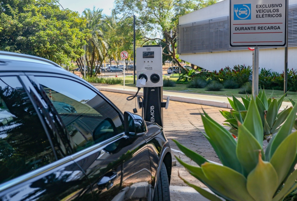 A imagem mostra uma estação de recarga para veículos elétricos em um estacionamento ao ar livre. Um carro preto está conectado à estação por meio de um cabo de carregamento. O equipamento possui uma tela de exibição e duas entradas, com a marca WEMOB visível. Ao lado, há uma placa indicando: “EXCLUSIVO VEÍCULOS ELÉTRICOS DURANTE RECARGA”, ou seja, a vaga é reservada apenas para carros elétricos enquanto estão carregando.