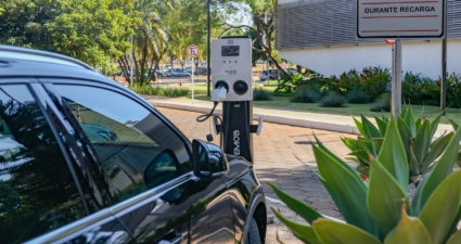 A imagem mostra uma estação de recarga para veículos elétricos em um estacionamento ao ar livre. Um carro preto está conectado à estação por meio de um cabo de carregamento. O equipamento possui uma tela de exibição e duas entradas, com a marca WEMOB visível. Ao lado, há uma placa indicando: “EXCLUSIVO VEÍCULOS ELÉTRICOS DURANTE RECARGA”, ou seja, a vaga é reservada apenas para carros elétricos enquanto estão carregando.