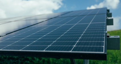 A imagem mostra um conjunto de painéis solares instalados ao ar livre. Os módulos fotovoltaicos estão organizados em formato retangular, formando uma grade sobre uma estrutura metálica. Ao fundo, vê-se um céu parcialmente nublado e uma área de grama verde, indicando que os painéis estão em um espaço aberto — possivelmente um parque solar ou uma instalação de energia renovável. A cena transmite claramente a ideia de geração de energia limpa e sustentável, aproveitando a luz do sol para produzir eletricidade e contribuir para a preservação ambiental.