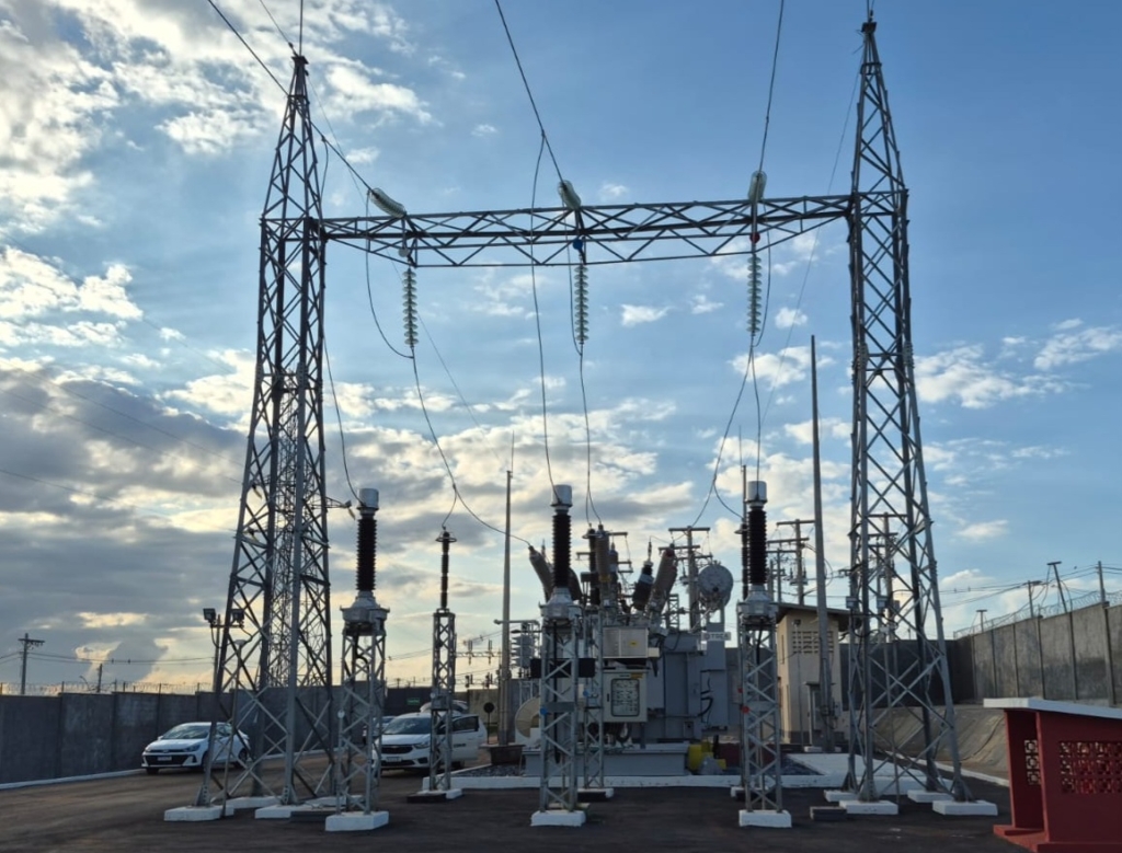 imagem mostra uma subestação elétrica.
Estruturas metálicas altas sustentam linhas de transmissão de alta tensão e isoladores.
No centro, há um grande transformador de potência, cercado por disjuntores, buchas e outros equipamentos usados para regular e distribuir a energia elétrica.
O espaço é delimitado por um muro de concreto, e alguns carros estão estacionados próximos, indicando que é uma área de acesso técnico.
O céu está parcialmente nublado, com a luz do sol iluminando a cena, o que dá contraste às estruturas metálicas.
Essa infraestrutura é essencial para o sistema de transmissão e distribuição de energia, garantindo que a eletricidade gerada em usinas chegue de forma segura às cidades e indústrias.