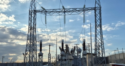 imagem mostra uma subestação elétrica. Estruturas metálicas altas sustentam linhas de transmissão de alta tensão e isoladores. No centro, há um grande transformador de potência, cercado por disjuntores, buchas e outros equipamentos usados para regular e distribuir a energia elétrica. O espaço é delimitado por um muro de concreto, e alguns carros estão estacionados próximos, indicando que é uma área de acesso técnico. O céu está parcialmente nublado, com a luz do sol iluminando a cena, o que dá contraste às estruturas metálicas. Essa infraestrutura é essencial para o sistema de transmissão e distribuição de energia, garantindo que a eletricidade gerada em usinas chegue de forma segura às cidades e indústrias.