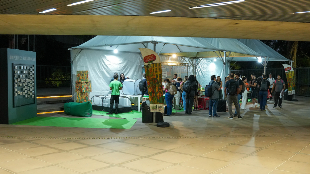 A imagem mostra um evento comunitário realizado ao ar livre, sob uma grande tenda iluminada. O espaço tem um clima descontraído, com piso verde e várias almofadas estilo “beanbag” espalhadas, convidando as pessoas a se sentarem e interagirem. À esquerda, há um painel vertical com o título “DISPONÍVEIS PARA ADOÇÃO”, onde estão fixadas diversas pequenas fotos ou cartões, sugerindo que se trata de uma iniciativa de adoção — possivelmente de animais ou livros. Em volta, vemos mesas e expositores, com pessoas conversando e participando das atividades. O ambiente é colorido e festivo, reforçado por banners e cartazes ilustrados, transmitindo uma atmosfera de convivência social e de conscientização. A cena parece capturar um momento de encontro comunitário voltado para promover adoção e engajamento social.