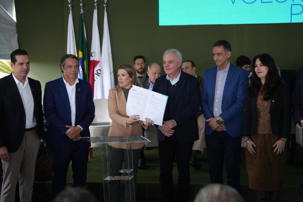 A imagem mostra um grupo de pessoas reunidas em um ambiente formal, provavelmente durante uma cerimônia ou evento oficial. No centro, uma pessoa segura um documento com texto e assinaturas visíveis, indicando a apresentação ou assinatura de um acordo ou parceria.
Ao fundo, há várias bandeiras expostas — entre elas, a do Brasil — reforçando o caráter institucional do momento. O cenário inclui uma parede verde e uma tela grande com texto parcialmente visível, sugerindo que se trata de um espaço preparado para a ocasião.
A cena transmite a ideia de reconhecimento oficial ou colaboração entre representantes, destacando a solenidade e a importância do encontro.