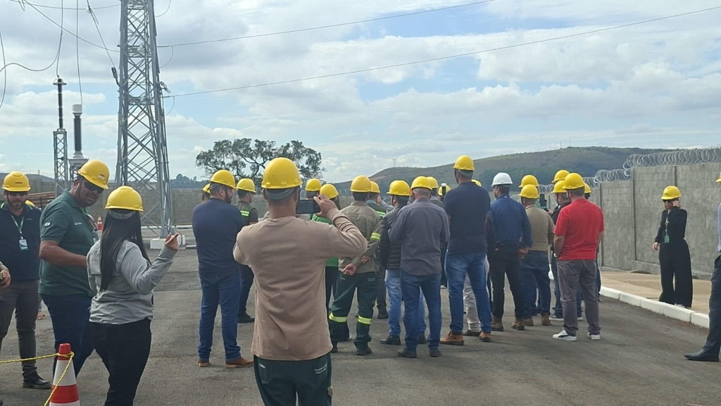 A imagem mostra um grupo de pessoas reunidas em um ambiente externo, próximo a uma estrutura de transmissão elétrica. Todos estão usando capacetes de segurança — alguns brancos, outros amarelos — o que indica que se trata de uma atividade técnica ou de inspeção.

O cenário inclui torres metálicas, cabos e isoladores, reforçando que é um local ligado à infraestrutura elétrica. A maioria veste uniformes de trabalho ou roupas casuais adequadas para campo, e uma pessoa aparece registrando o momento com um celular. Ao fundo, há colinas, árvores e um céu parcialmente nublado, compondo um contraste entre natureza e tecnologia.

A cena transmite a ideia de um treinamento ou vistoria profissional, destacando práticas de segurança e trabalho em equipe em um ambiente industrial.