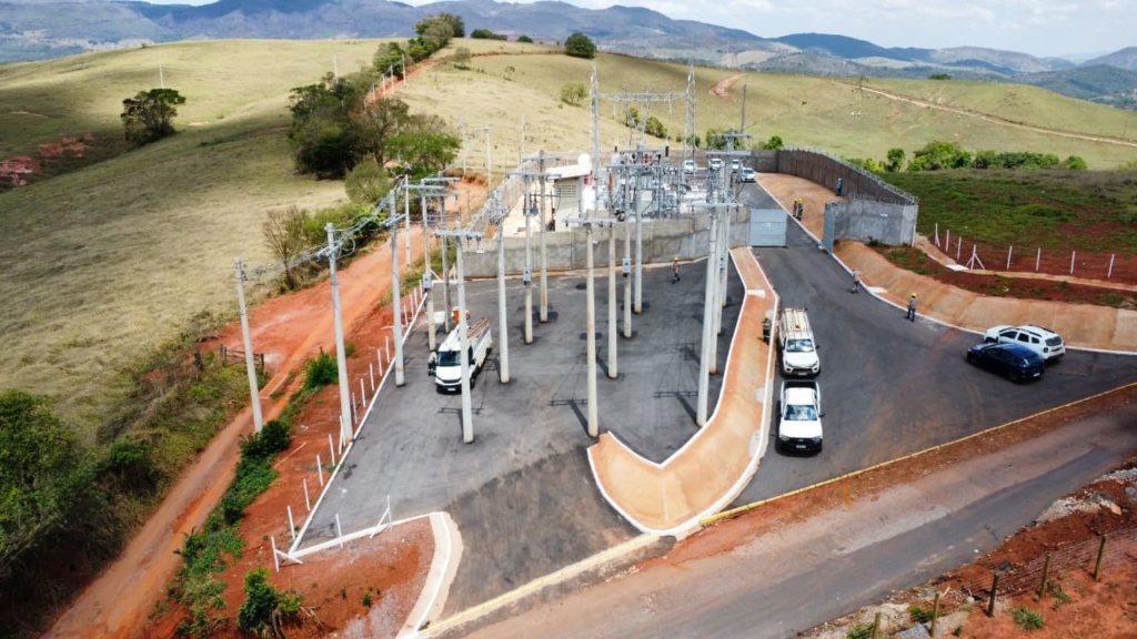A imagem mostra uma subestação elétrica localizada em uma área rural, cercada por colinas e campos abertos. Dentro do perímetro cercado, há diversos postes de concreto altos sustentando linhas de transmissão e equipamentos como transformadores e disjuntores.

Alguns veículos utilitários estão estacionados na área pavimentada, e trabalhadores com equipamentos de segurança aparecem próximos às estruturas, indicando manutenção ou operação em andamento. O solo ao redor é avermelhado, com vegetação esparsa, e uma estrada pavimentada dá acesso direto à instalação.

A cena transmite a importância dessa infraestrutura para a distribuição de energia elétrica, mostrando como a eletricidade é gerida e transmitida mesmo em regiões remotas ou semi-rurais.