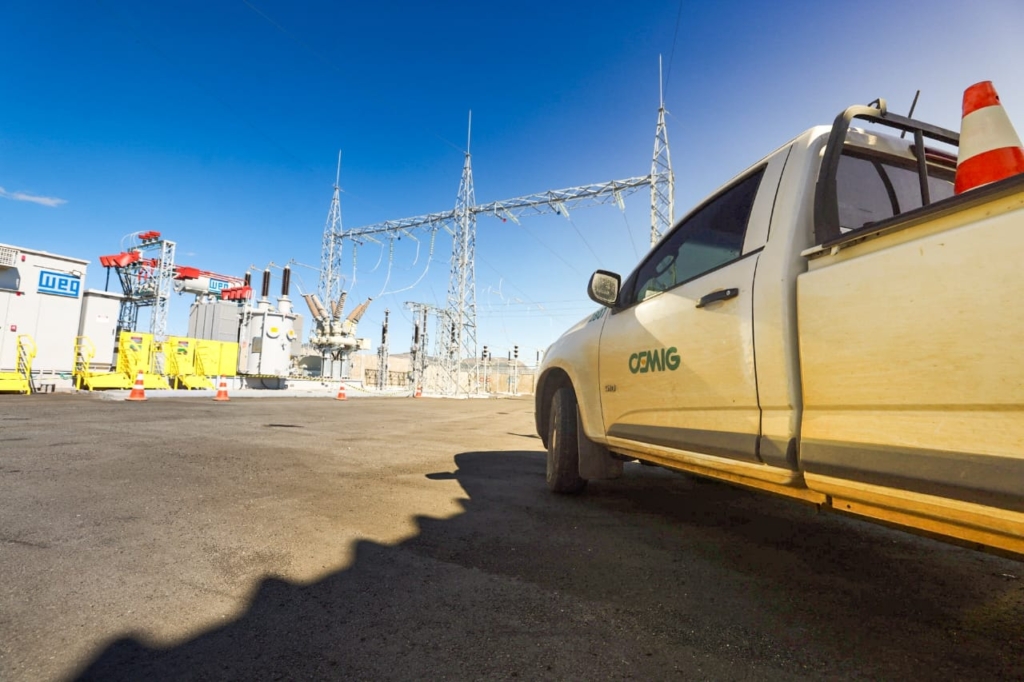 A imagem mostra uma subestação elétrica em operação durante o dia, sob um céu azul e claro. No primeiro plano, há uma caminhonete branca com o logotipo CEMIG na porta, utilizada para serviços de manutenção. Na caçamba, vê-se um cone de sinalização laranja.

Ao redor, estão dispostos diversos equipamentos de alta tensão, como transformadores, disjuntores e estruturas de transmissão com linhas aéreas. Também aparecem barreiras de segurança amarelas e vários cones laranja posicionados para delimitar áreas de trabalho, reforçando as práticas de segurança no local.

O cenário evidencia a importância dessa infraestrutura para a distribuição de energia elétrica, mostrando tanto os equipamentos essenciais quanto os recursos de manutenção e segurança empregados.