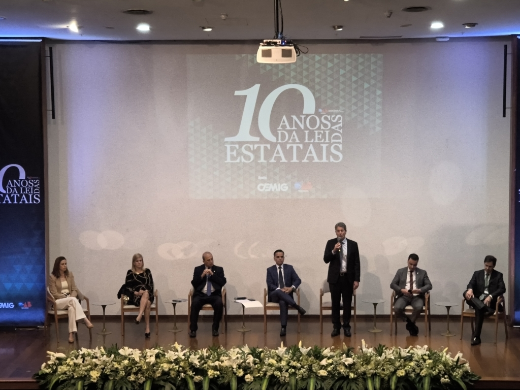 A imagem mostra um evento formal realizado em um auditório. No palco, há sete pessoas sentadas e uma em pé, falando ao microfone. A frente do palco está decorada com um arranjo de flores, reforçando o caráter solene da ocasião.

Ao fundo, um grande telão exibe a frase “10 ANOS DA LEI DAS ESTATAIS”, acompanhada dos logotipos da CEMIG e da OAB. À esquerda, um banner vertical traz o mesmo texto, reforçando o tema central da conferência.

O cenário transmite a ideia de uma comemoração institucional, destacando a importância da lei das estatais no Brasil e o papel das entidades envolvidas.