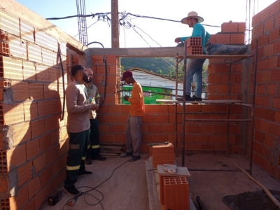 A imagem mostra um canteiro de obras em fase inicial de construção. As paredes de tijolos vermelhos estão sendo erguidas e ainda não há telhado instalado. Quatro pessoas trabalham no local: uma delas está sobre um andaime, usando um chapéu de aba larga e manuseando tijolos, enquanto as outras três permanecem no chão, aparentemente discutindo ou avaliando o andamento da obra. No chão, há pilhas de tijolos, ferramentas de construção e cabos elétricos espalhados. Ao fundo, vê-se um prédio verde e algumas colinas, indicando que o cenário é ao ar livre. A cena transmite a ideia de trabalho em equipe e progresso na construção, destacando o esforço manual e a coordenação necessários para levantar uma estrutura.