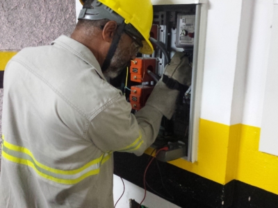 A imagem mostra um profissional realizando trabalho elétrico em um painel de distribuição montado na parede. Ele está usando equipamentos de segurança: capacete amarelo, óculos de proteção e uniforme cinza claro com faixas refletivas. O painel contém diversos componentes, como fios, disjuntores e chaves, e o trabalhador parece estar inspecionando ou ajustando a fiação com ferramentas. O ambiente transmite a ideia de manutenção técnica e práticas seguras, reforçadas pela faixa amarela e preta pintada na parede atrás dele, que sugere uma área de risco controlado. É uma cena que evidencia tanto a segurança no trabalho quanto a expertise técnica envolvida nesse tipo de atividade.