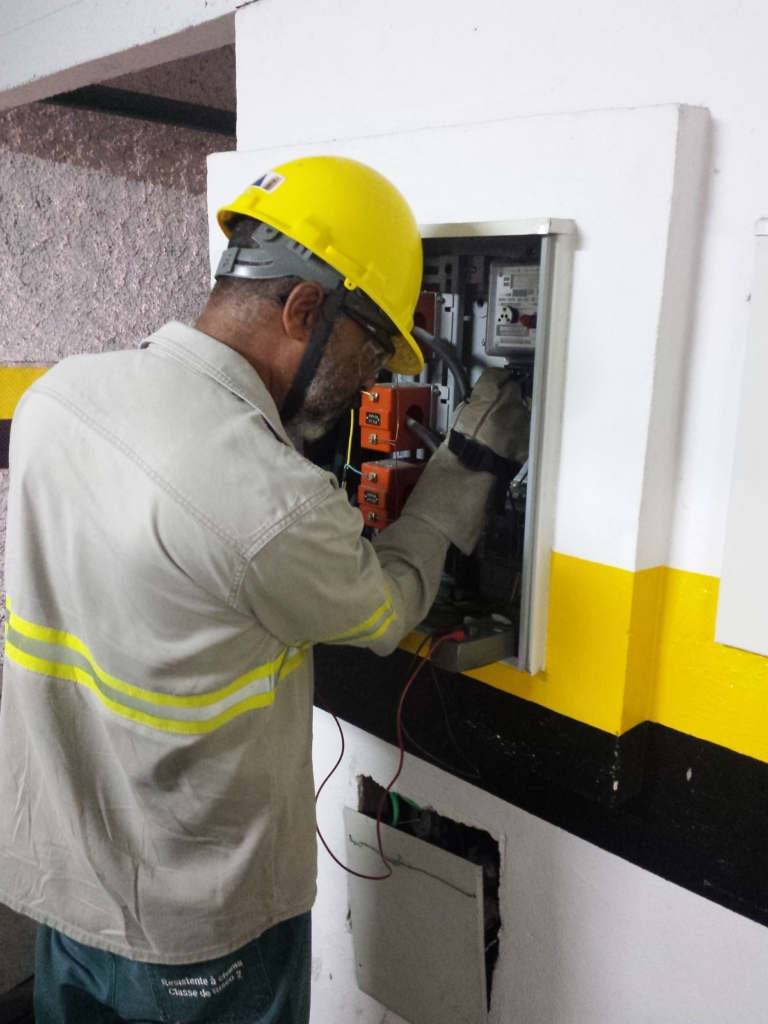 A imagem mostra um profissional realizando trabalho elétrico em um painel de distribuição montado na parede. Ele está usando equipamentos de segurança: capacete amarelo, óculos de proteção e uniforme cinza claro com faixas refletivas. O painel contém diversos componentes, como fios, disjuntores e chaves, e o trabalhador parece estar inspecionando ou ajustando a fiação com ferramentas.

O ambiente transmite a ideia de manutenção técnica e práticas seguras, reforçadas pela faixa amarela e preta pintada na parede atrás dele, que sugere uma área de risco controlado. É uma cena que evidencia tanto a segurança no trabalho quanto a expertise técnica envolvida nesse tipo de atividade.