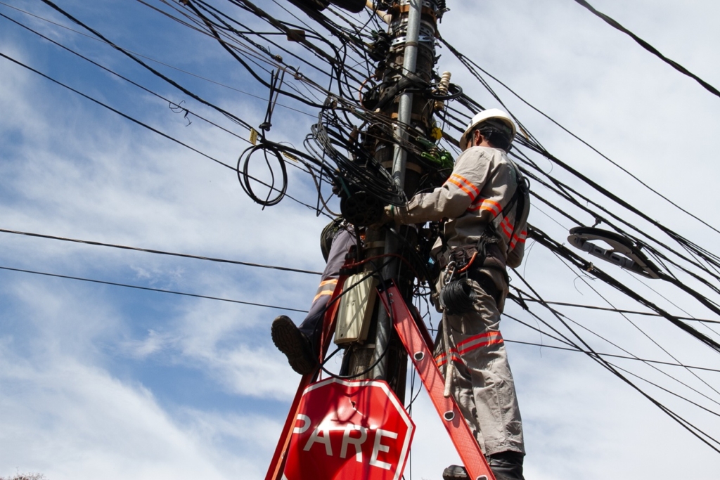 A imagem mostra um trabalhador de utilidade pública usando equipamentos de segurança e capacete, posicionado em uma escada vermelha apoiada em um poste de energia e comunicação. O poste está repleto de cabos elétricos e de telecomunicação, muitos deles visivelmente emaranhados, o que transmite a ideia de complexidade e desafio técnico. O trabalhador parece estar realizando manutenção ou instalação nos fios.

Na parte inferior do poste há uma placa de trânsito vermelha com a palavra “PARE”, indicando “STOP” em espanhol/português. O céu ao fundo está parcialmente nublado, com algumas áreas de azul visíveis, criando contraste com a cena urbana.

Esse tipo de imagem evidencia tanto a dificuldade da infraestrutura elétrica e de comunicação quanto a importância da segurança no trabalho em altura.