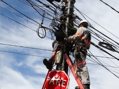 A imagem mostra um trabalhador de utilidade pública usando equipamentos de segurança e capacete, posicionado em uma escada vermelha apoiada em um poste de energia e comunicação. O poste está repleto de cabos elétricos e de telecomunicação, muitos deles visivelmente emaranhados, o que transmite a ideia de complexidade e desafio técnico. O trabalhador parece estar realizando manutenção ou instalação nos fios. Na parte inferior do poste há uma placa de trânsito vermelha com a palavra “PARE”, indicando “STOP” em espanhol/português. O céu ao fundo está parcialmente nublado, com algumas áreas de azul visíveis, criando contraste com a cena urbana. Esse tipo de imagem evidencia tanto a dificuldade da infraestrutura elétrica e de comunicação quanto a importância da segurança no trabalho em altura.